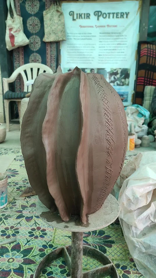 Unfinished ceramic or clay sculpture of a large petal or shell shape, in a workshop with various tools, fabrics, and a sign that reads 'Likir Pottery' in the background.