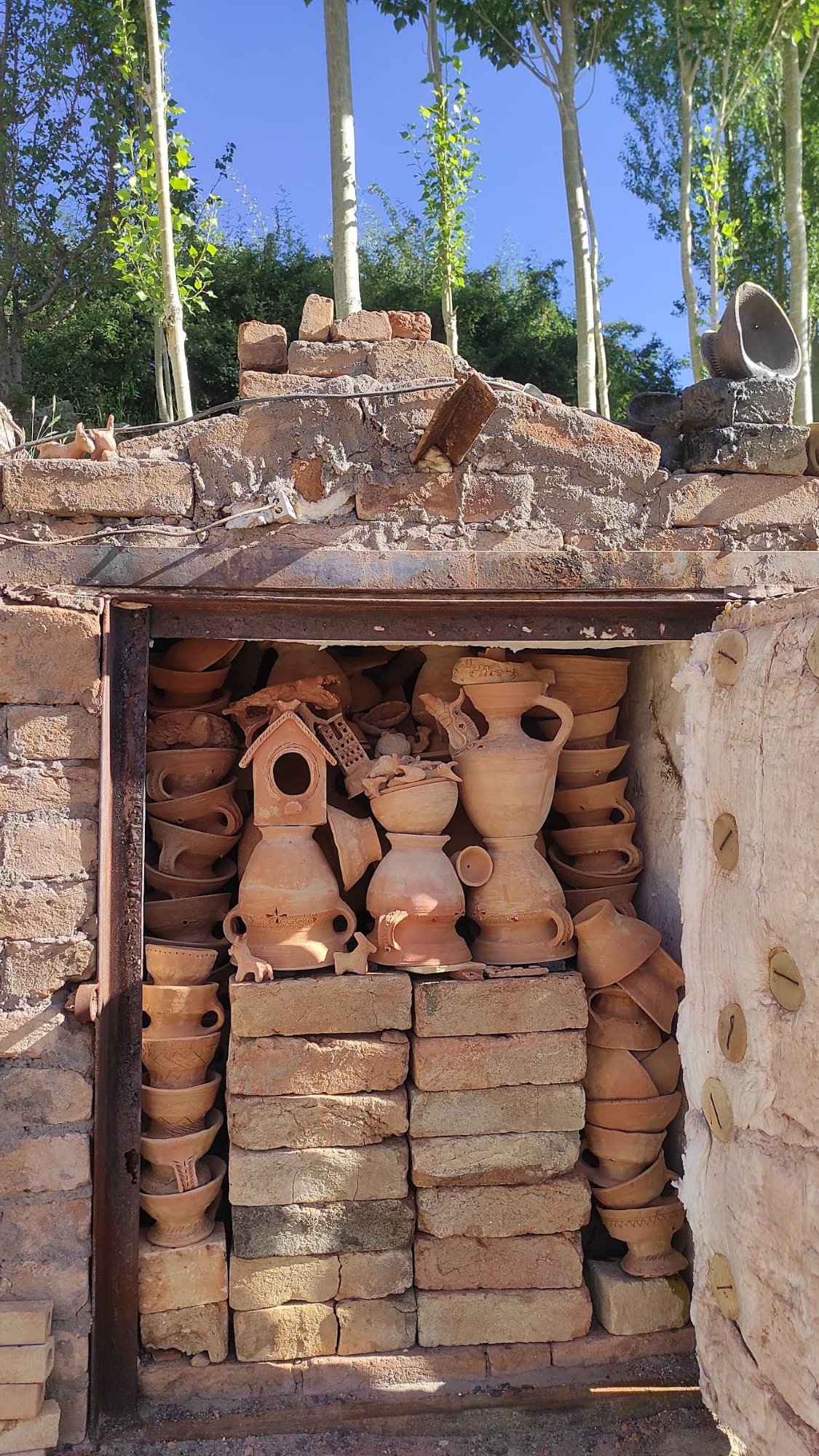 Shelves filled with various unfinished terracotta and clay pottery, such as vases, pots, and decorative items, stored in an outdoor brick oven or kiln.