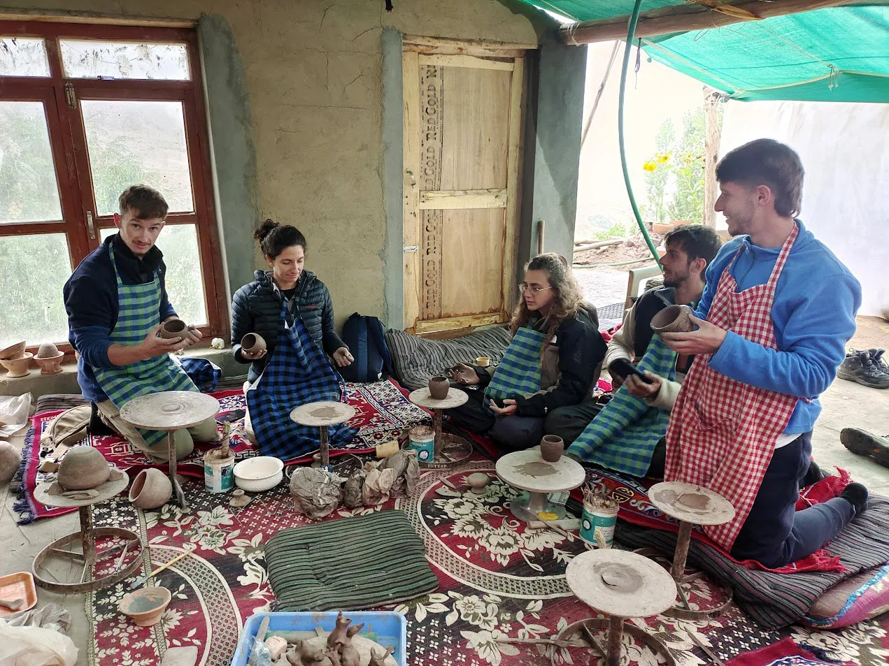 Group of people inside a rustic room, possibly a pottery workshop, sitting on cushions on a decorative rug, with pottery objects and tools around them, some holding and examining clay pots, engaging in pottery making.