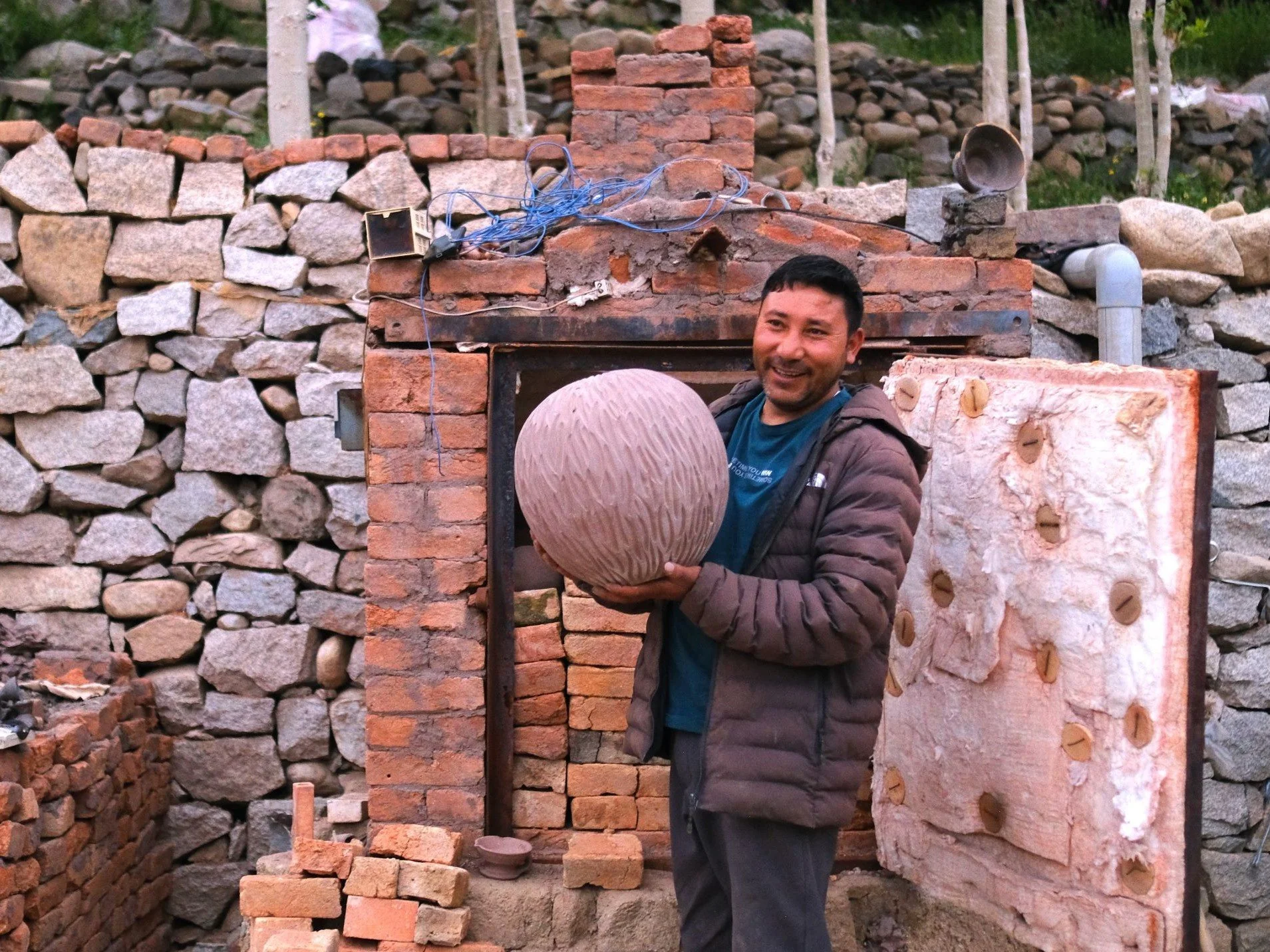 A man in a brown jacket smiling and holding a large clay pot in front of a brick gas kiln under construction, with stone walls and construction materials around.