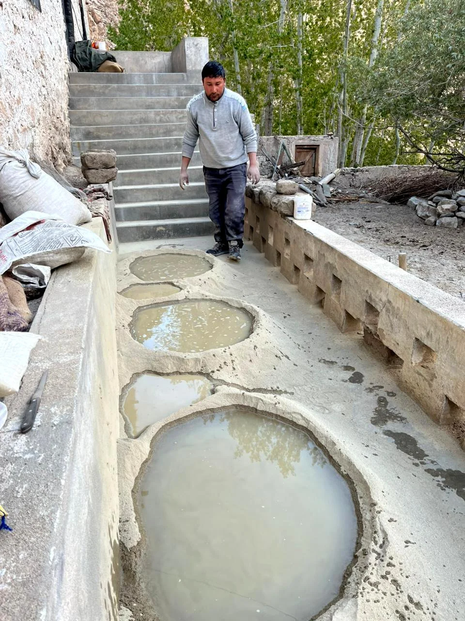 A man standing on a concrete pathway with four round, shallow pools filled with water at an outdoor construction site. The background includes a staircase and trees.