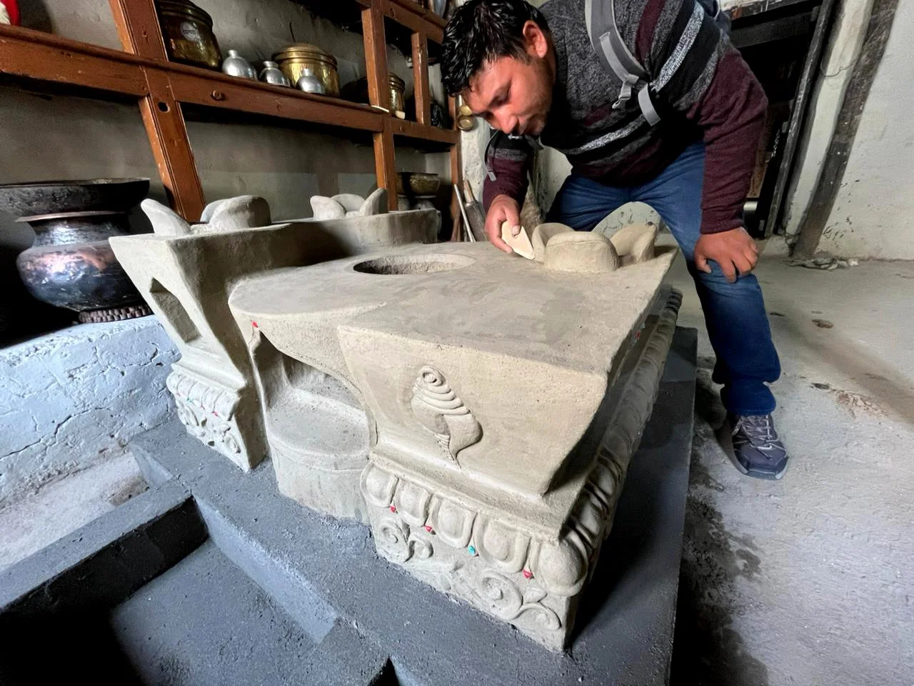 A man is carving details into an ancient clay stove in a workshop, surrounded by shelves with artifacts and pottery.