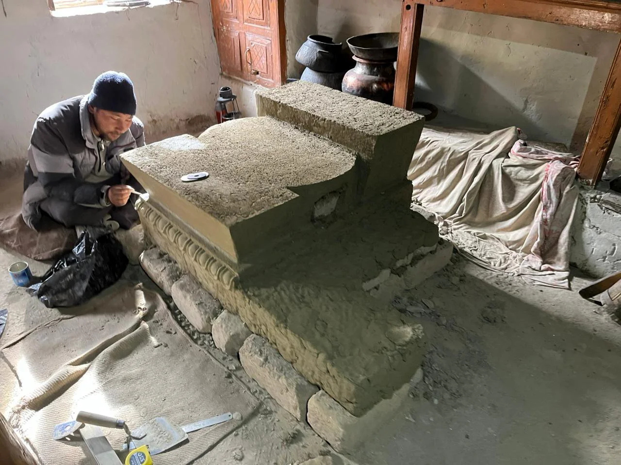 A person working on restoring or carving an ancient stone structure inside a rustic room, with tools and supplies nearby.