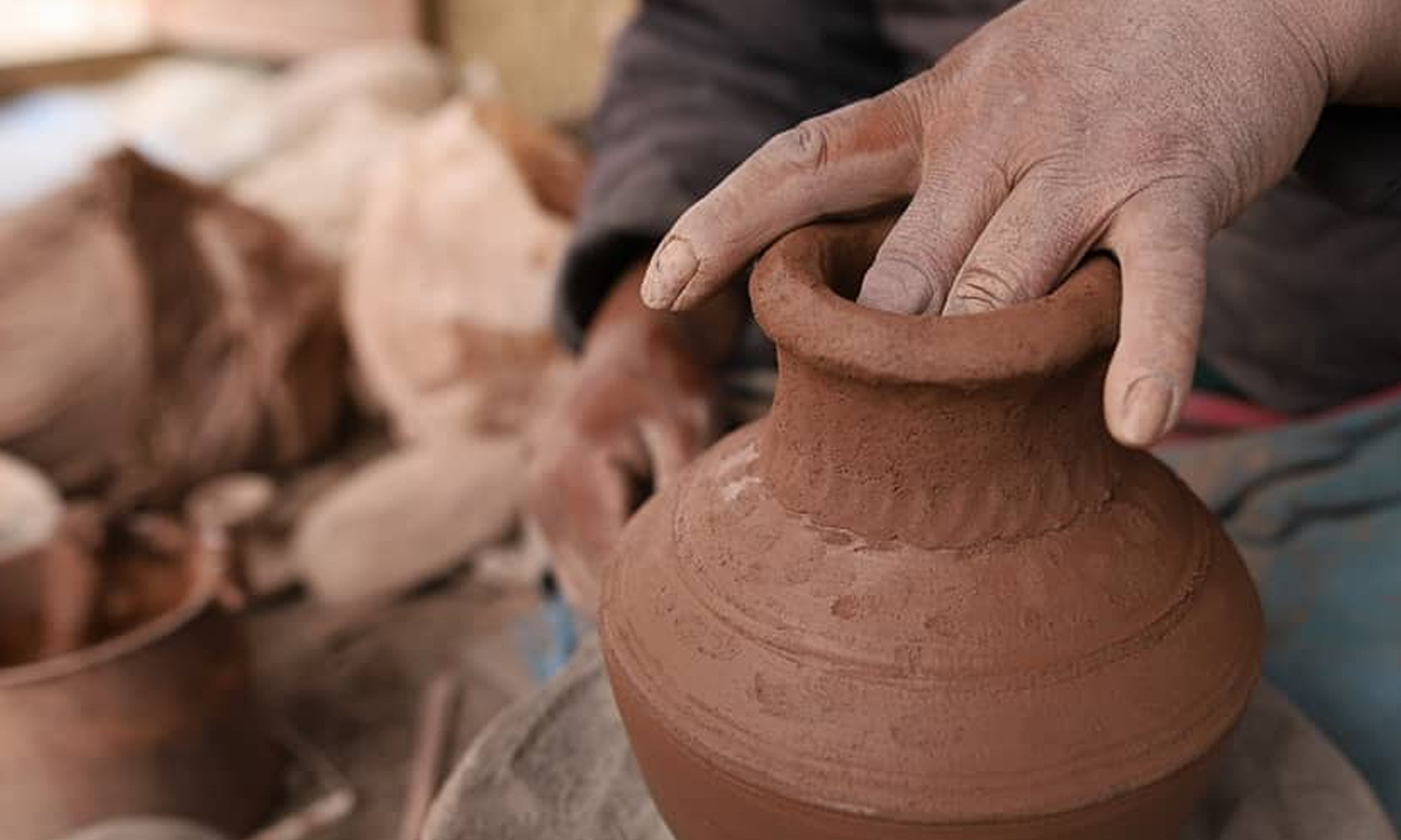 A person shaping a clay pot on a pottery wheel in a workshop, with a collection of finished or raw clay pieces in the background.