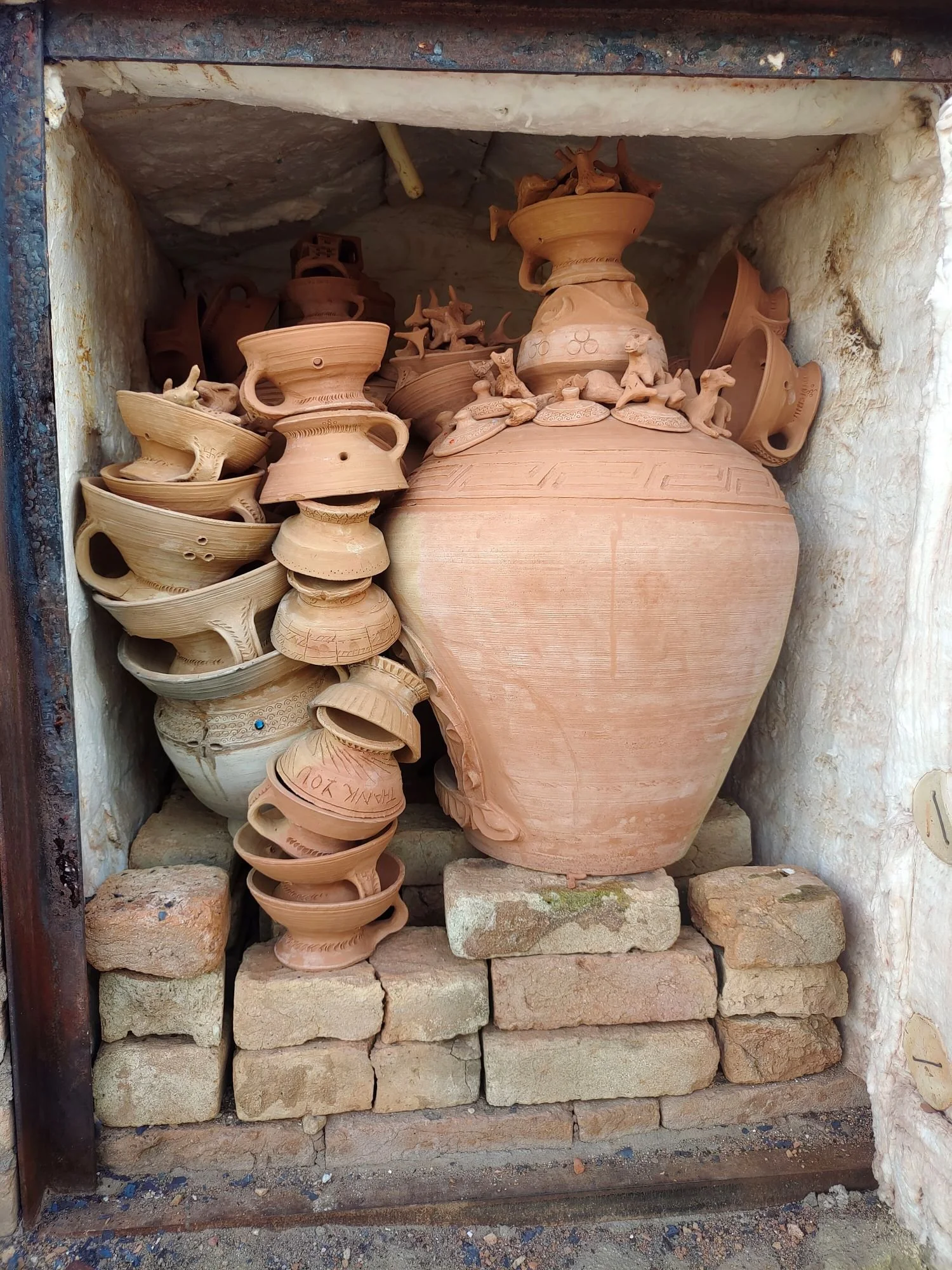 A collection of unglazed terracotta pottery including large vases, bowls, and small figurines of animals displayed on brick kiln.