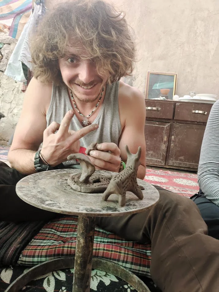A man with curly, messy hair, wearing a tank top and necklaces, is smiling and making a gesture with his hand while seated on the floor. He is holding a small sculpture of a dinosaur, positioned on a round, rustic table with a leopard print cushion. 