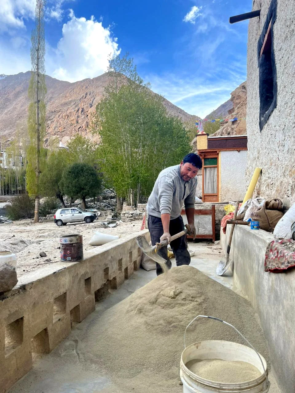 A man shovels sand or gravel at an outdoor construction site with mountains, trees, a car, and a stone building in the background.