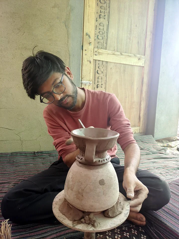 A young man with glasses and a beard, wearing a pink sweater, sitting cross-legged on a patterned mat inside a room. He is working on a clay pottery piece on a small turntable with a simple background, including a door and a textured wall.