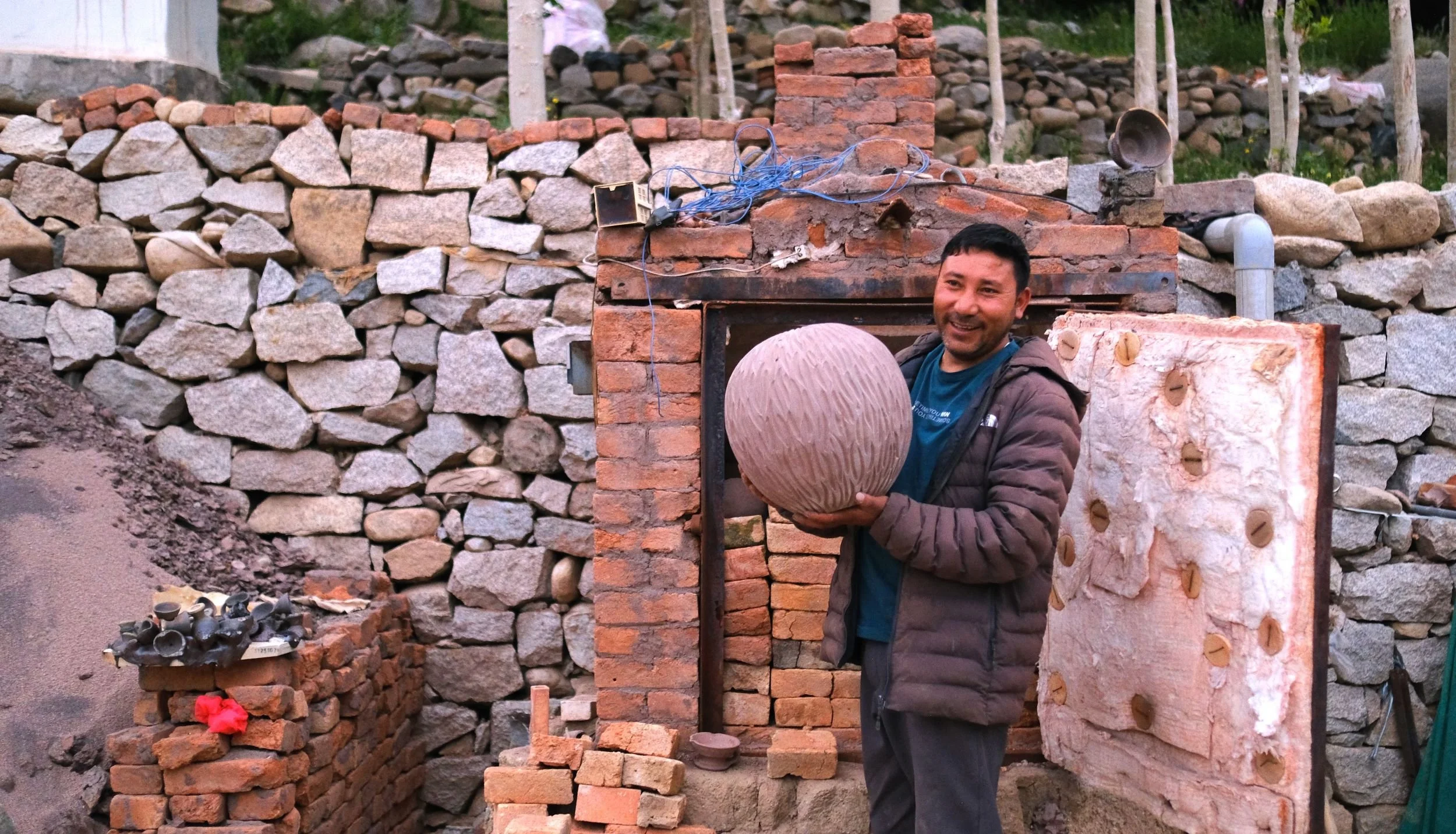 Rigzen Namgail smiling while holding a large, round piece of pottery in front of a brick and stone wall of a gas kiln.