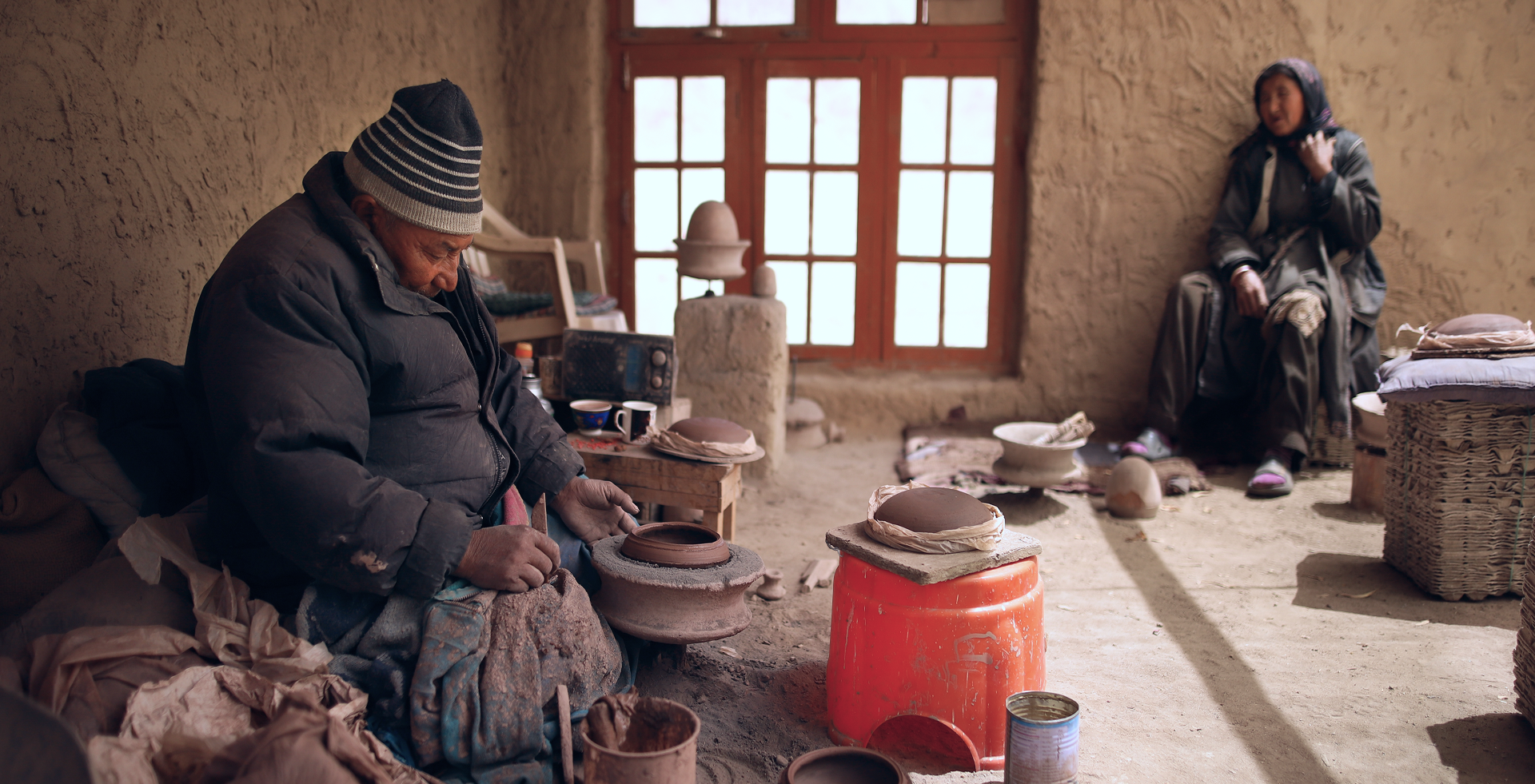 Abba'ley Lamchung Tsepail working on pottery inside a rustic studio with a Amma'ley sitting nearby. The room has a dirt floor, a window with multiple panes, and various pottery tools and supplies around.
