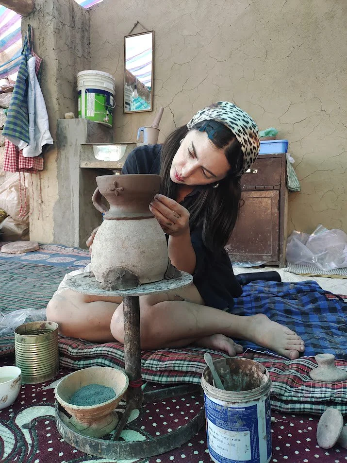A woman sits on the floor, shaping a clay pot in an outdoor workspace surrounded by pottery tools, containers, and fabric, with a rustic background and a small mirror on the wall.