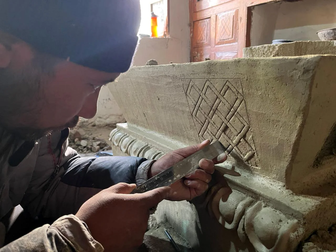A craftsman carving an intricate geometric pattern into a clay stove (thap) block using a chisel and hammer inside a workshop.