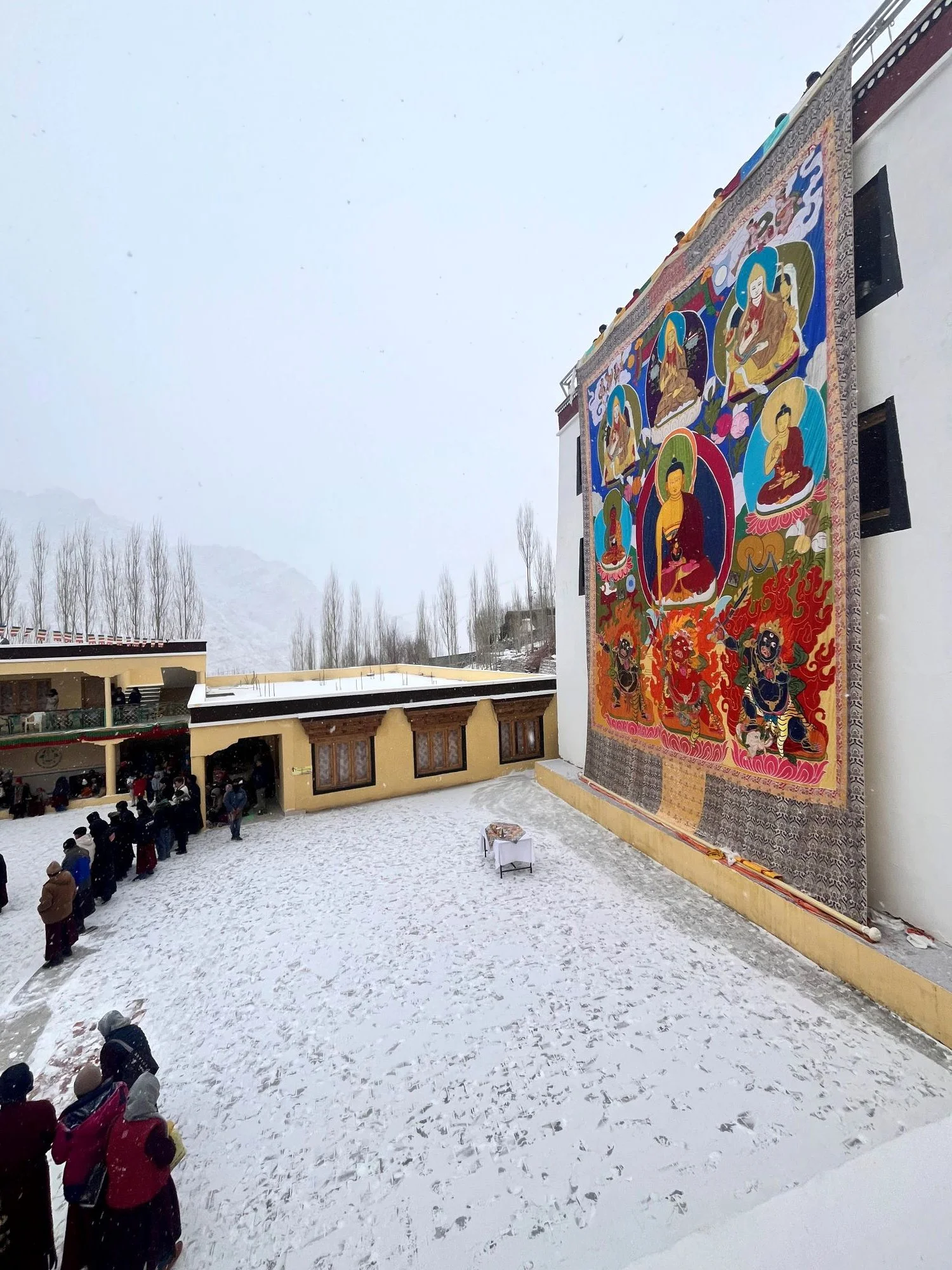 Outdoor courtyard with snow-covered ground and a large colorful Tibetan-style thangka hanging on a building wall. People are standing and walking in line beneath the thangka, with a small table in the center.