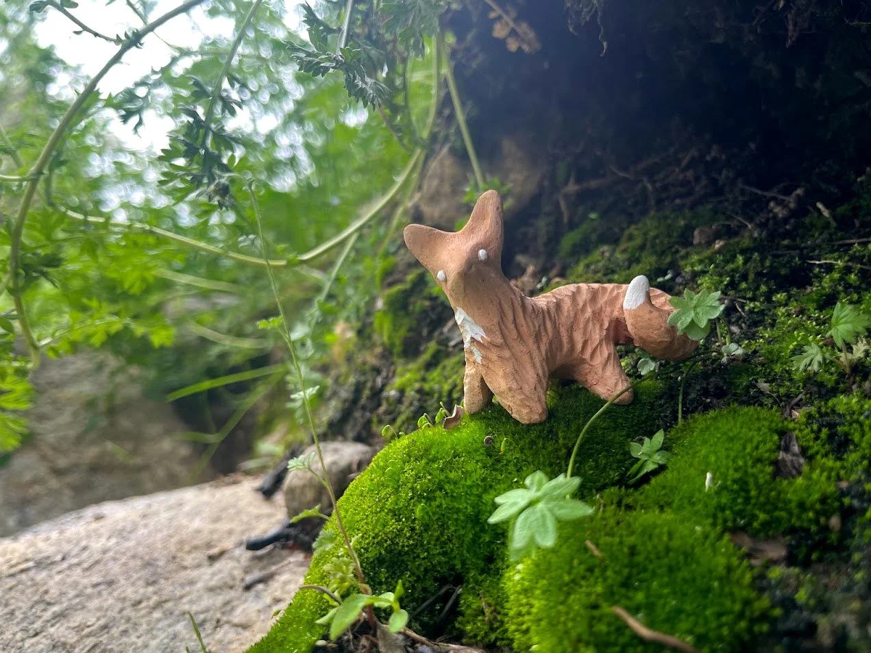 A small clay fox figurine with white accents on its face and tail, sitting on green moss among small plants and leaves, with blurred green foliage in the background.