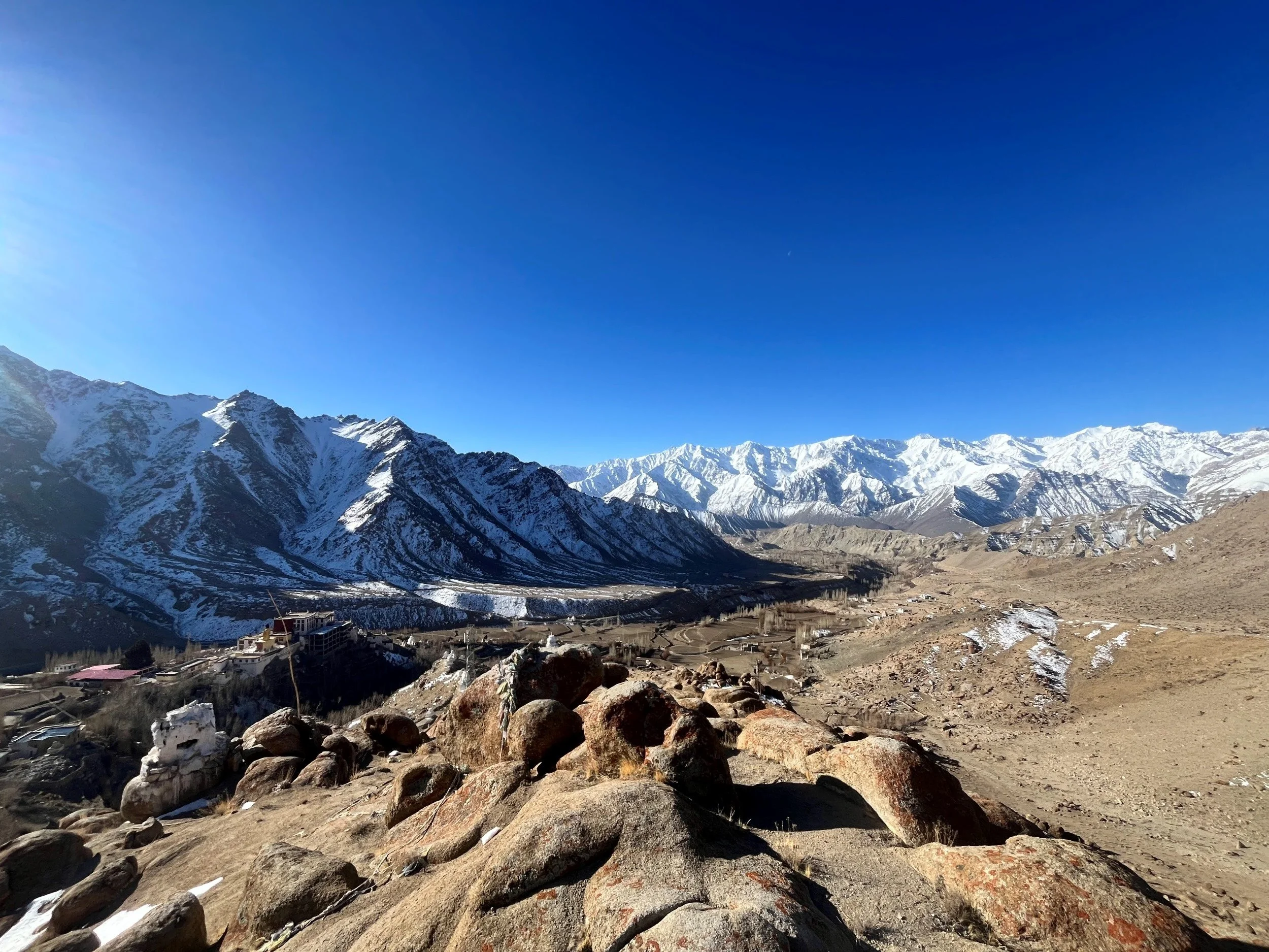 Snow-capped mountains under a clear blue sky with a few clouds, a small village - Likir and rugged terrain in the foreground.