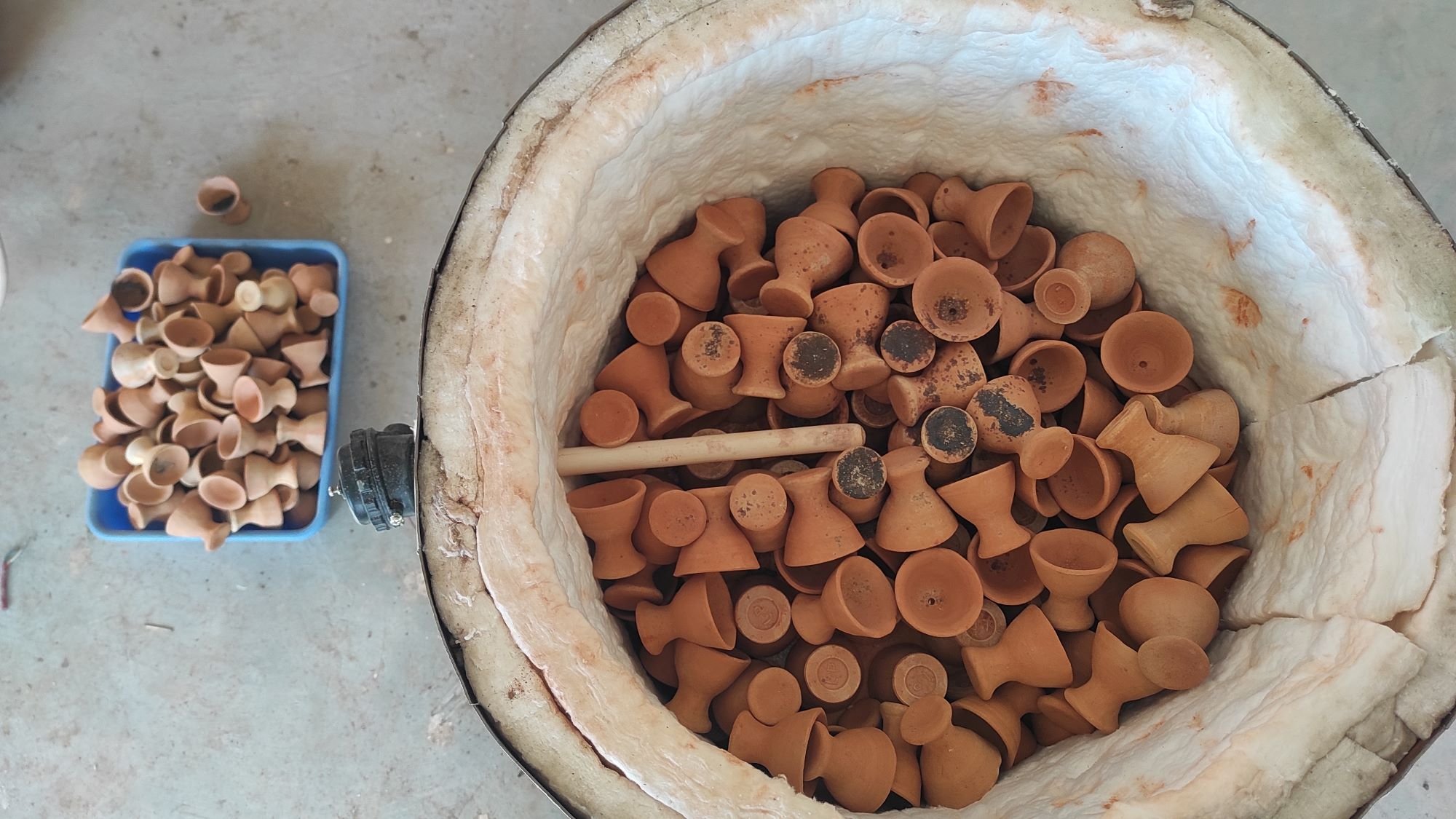 Clay pots inside a heated kiln, with a tray of more clay pots nearby on a concrete surface.
