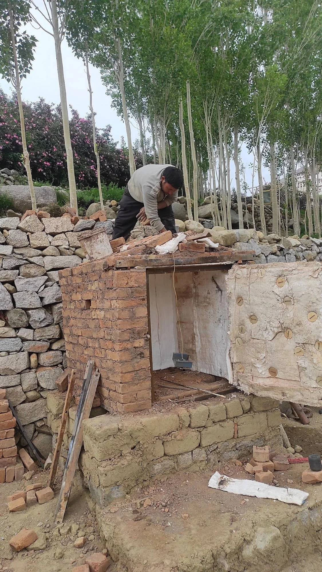 A man working on construction of an outdoor gas kiln with brick and stone, surrounded by trees and rocks.