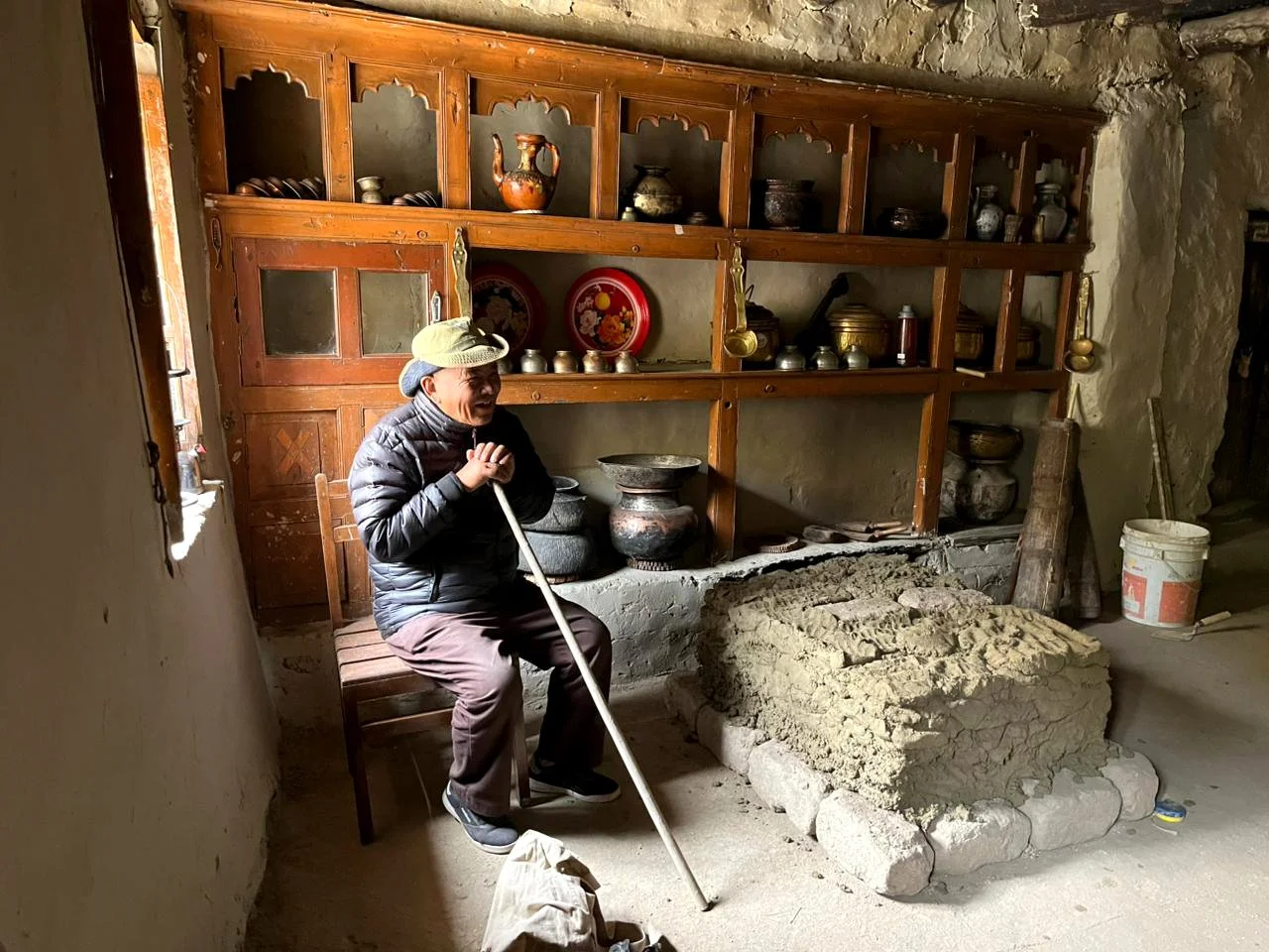 A person sitting on a wooden chair in a rustic room with a large stone oven or cooking area in front of him. The person is wearing a jacket, pants, a cap, and is holding a stick or pole. Behind him is a wooden shelf with various pots, bowls, and deco