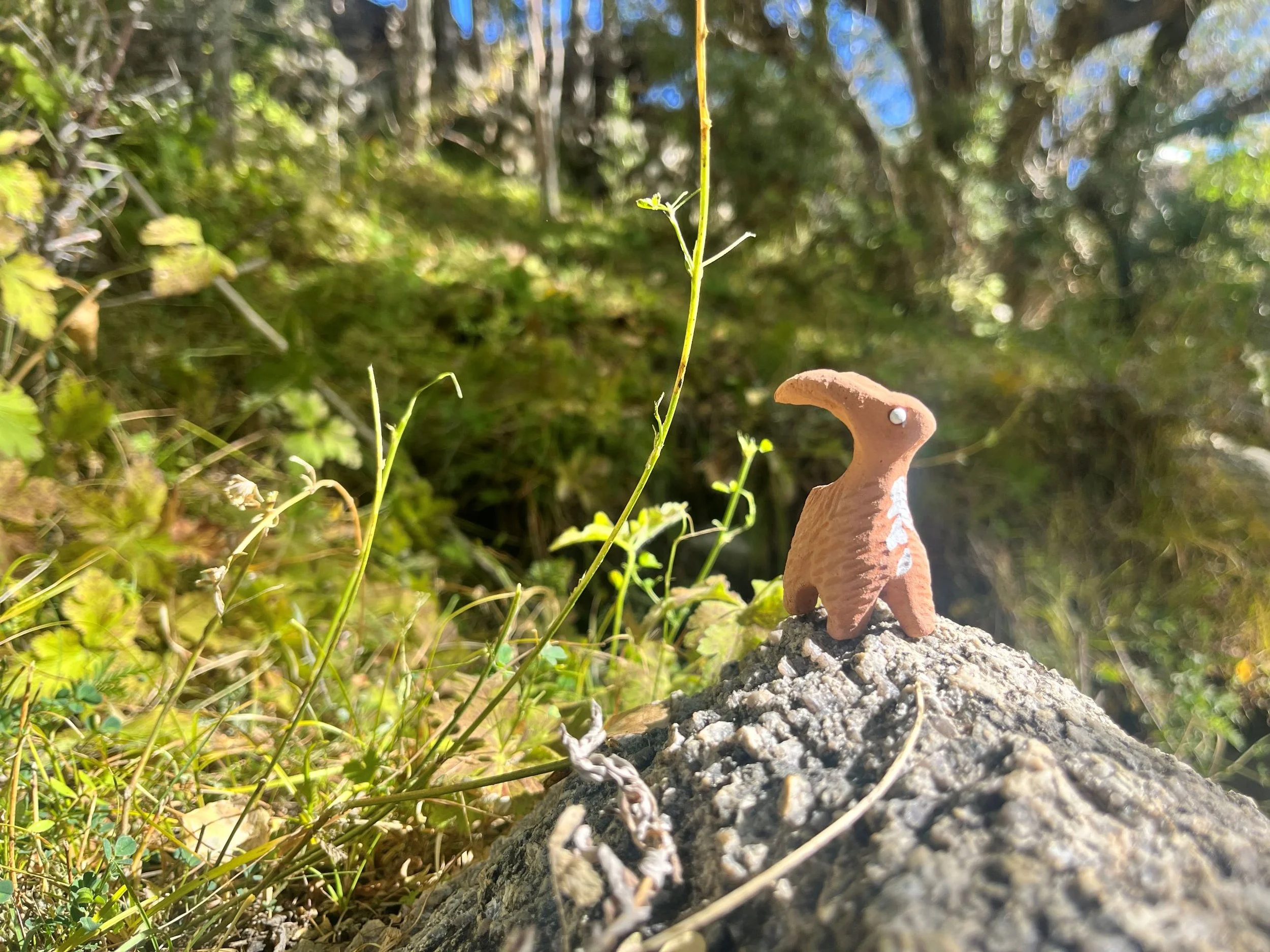 Close-up of a small terracotta figurine of a Ibex standing on gravel on the ground, with green plants and trees in the background.