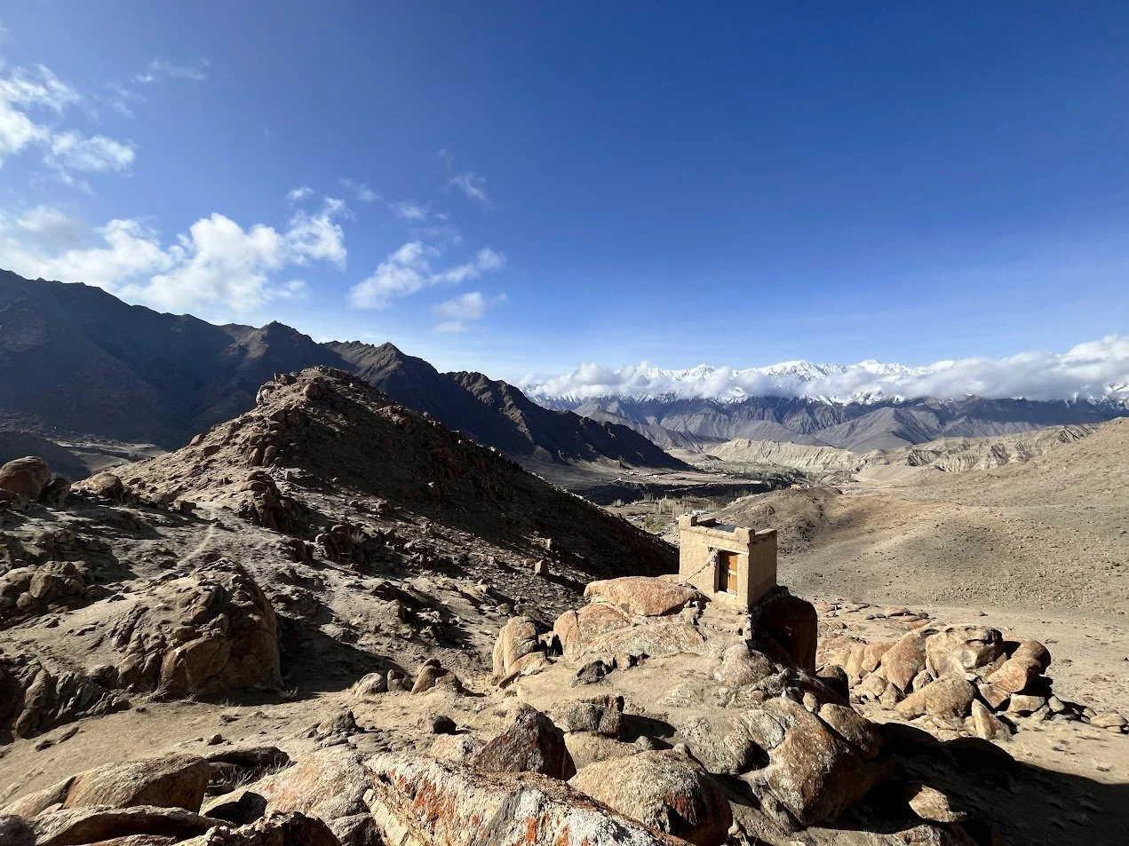 Barren rocky landscape with mountains in the distance under a clear blue sky, a small, old stone structure in the foreground.