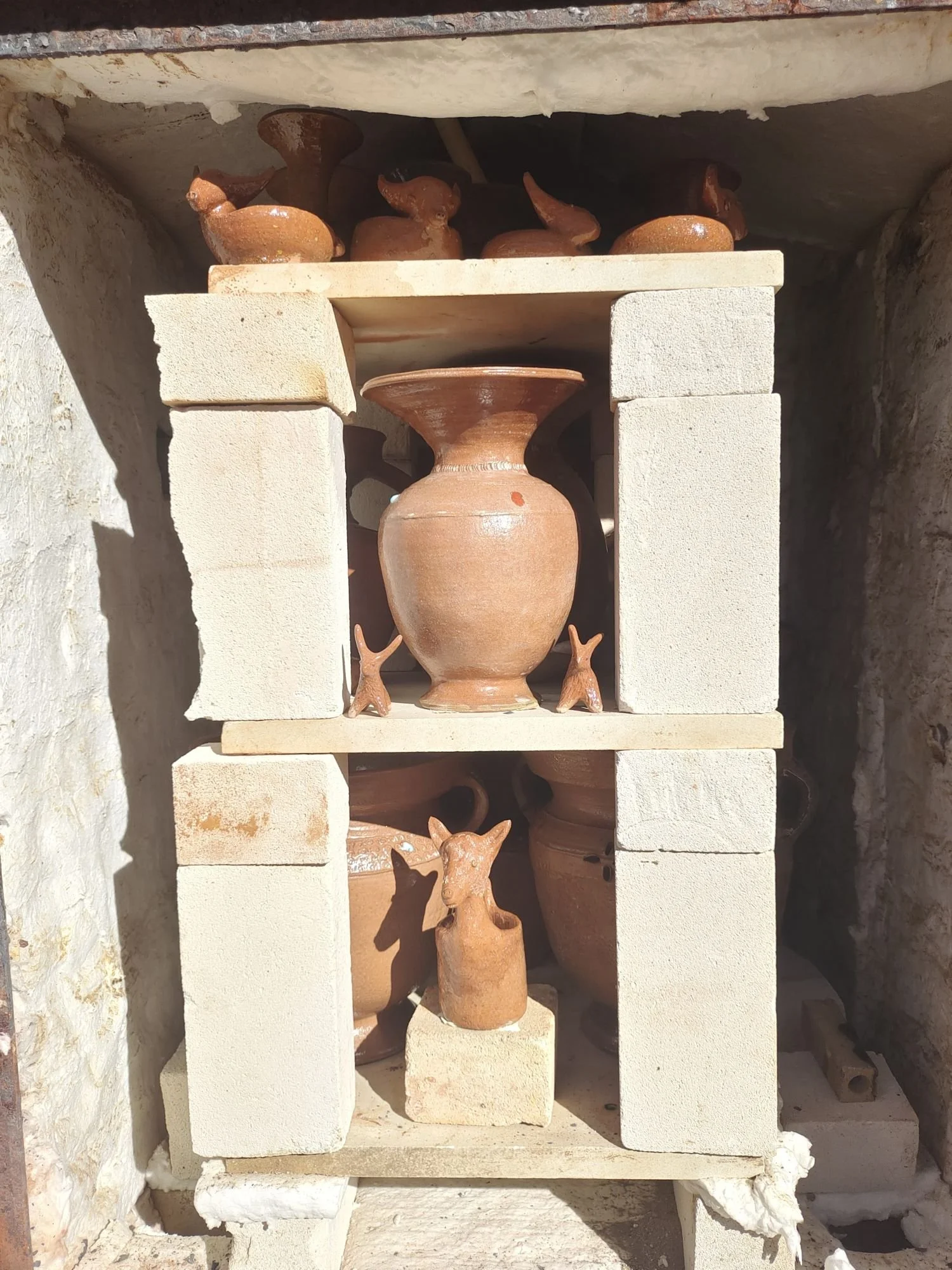 Display of ancientterracotta pottery and small animal figurines arranged on white brick shelves in a stone-walled kiln.