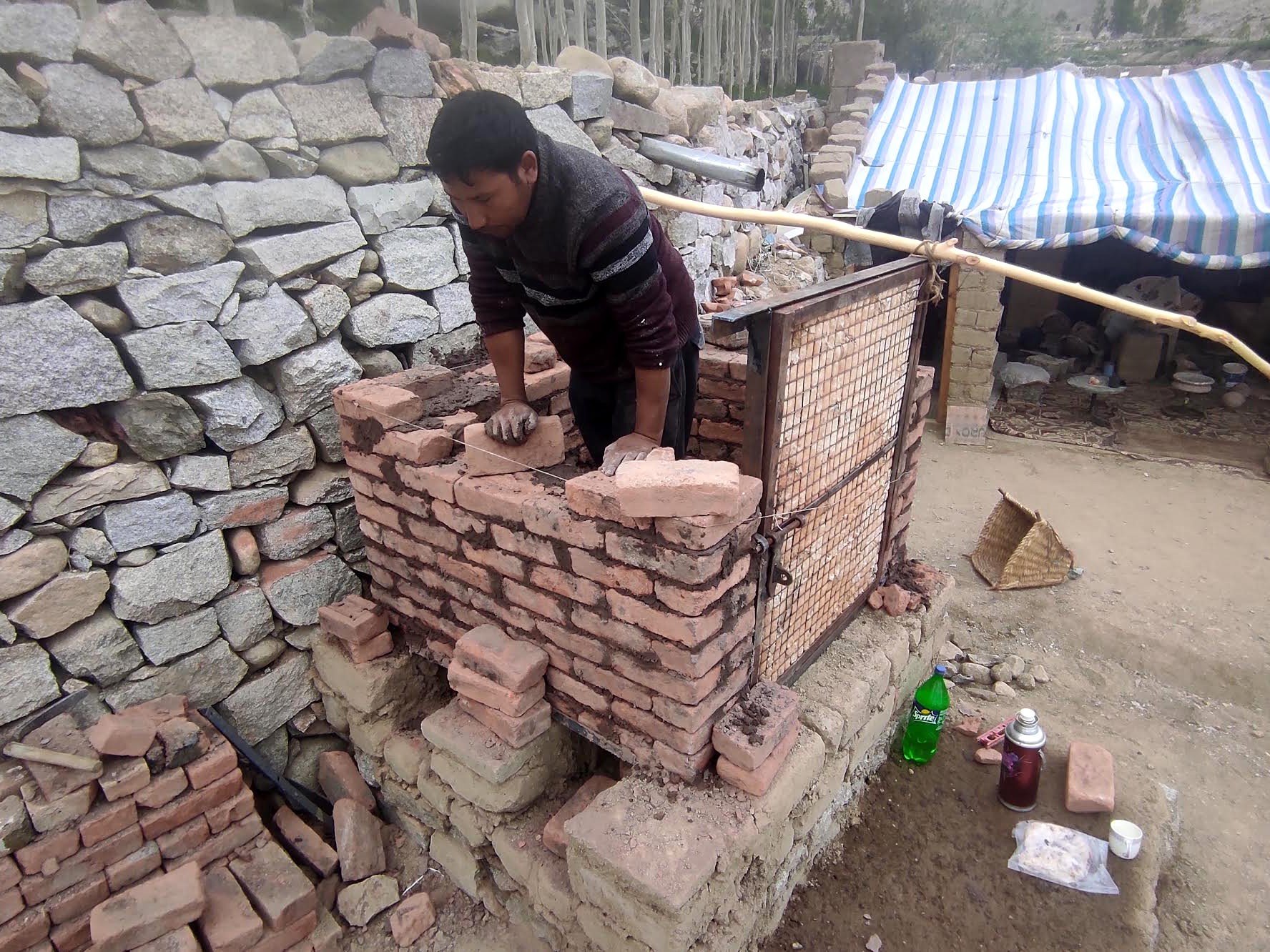 Rigzen Namgail constructing a brick wall on a kiln site with tools and supplies nearby.