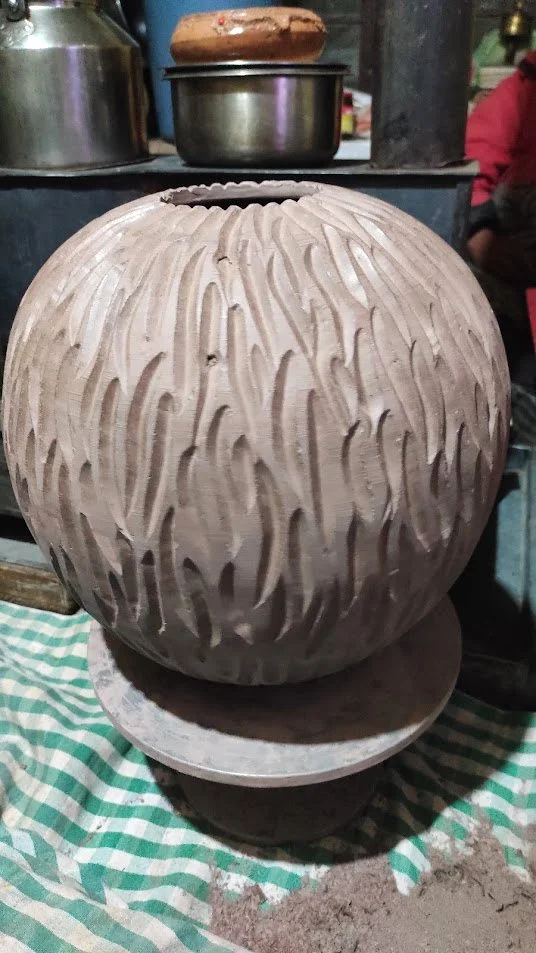 A large spherical ceramic pot with carved vertical lines, placed on a wheel in a pottery workshop.