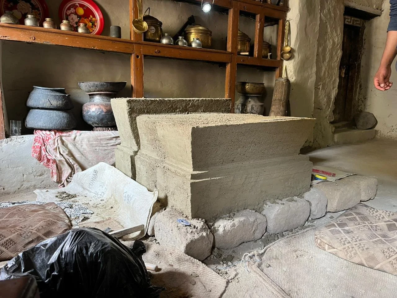 Interior of a rustic room with a large stone structure under construction, surrounded by cushions, canned goods, and kitchen utensils on wooden shelves.