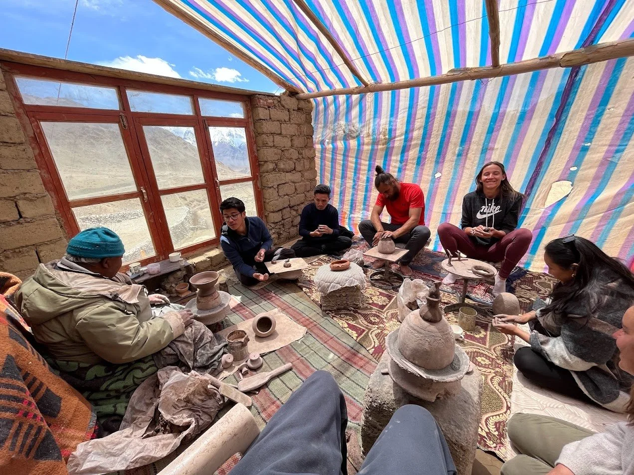 Group of people engaged in pottery making inside a rustic room with a window showing mountains outside, using pottery wheels and clay