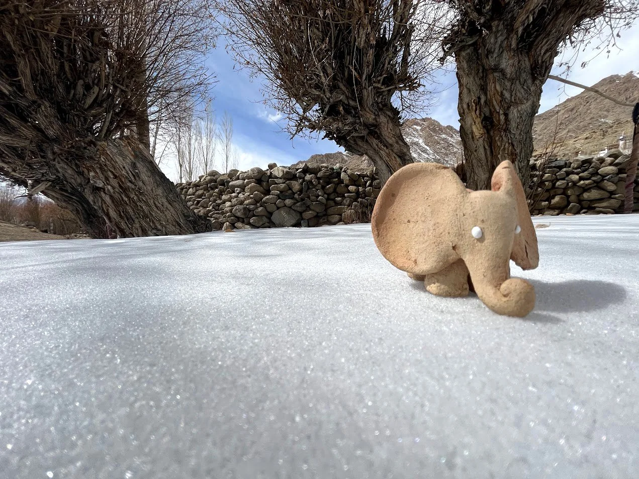 A small, beige clay elephant figurine with white eyes, placed on snow with trees and a stone wall in the background.