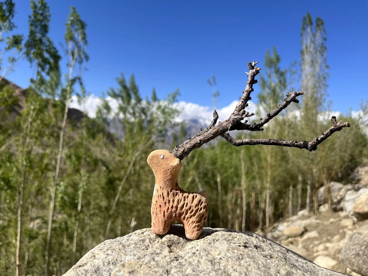 A small decorative sculpture of a dog with a branch for antlers, placed on a rock outdoors with green trees and a mountain in the background under a blue sky.