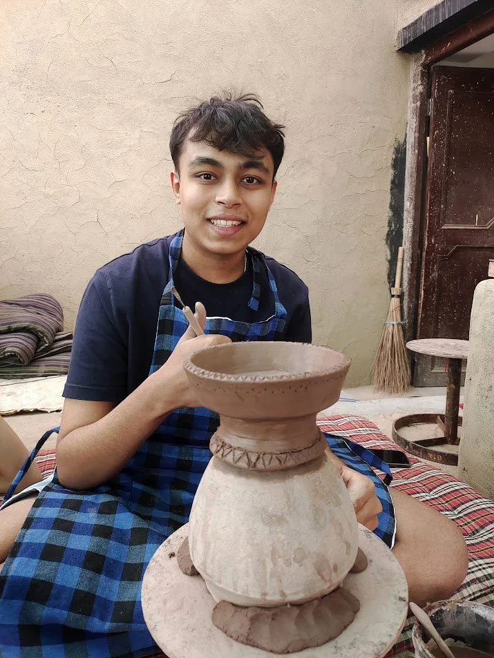 A young man with dark hair, wearing a black t-shirt and a blue checkered apron, sitting cross-legged on a patterned cloth, holding a clay bowl on a pottery wheel. The setting is an outdoor workshop with a beige wall, a broom, and a wooden door in the