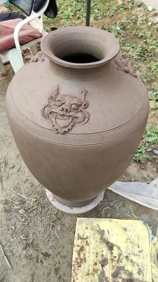 A large unfinished clay vase with a carved dragon design near the top, set outdoors on a wheel, surrounded by dirt and gardening items.
