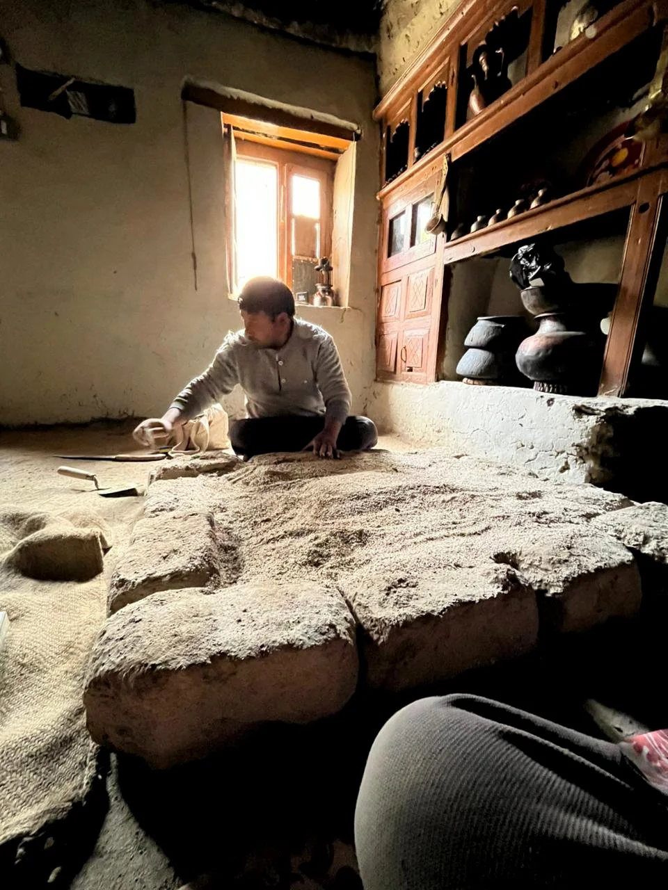 A person sitting on the floor of a rustic room, working on large stone slabs, with a window behind them letting in natural light, and wooden shelves with pottery and artifacts on the wall.