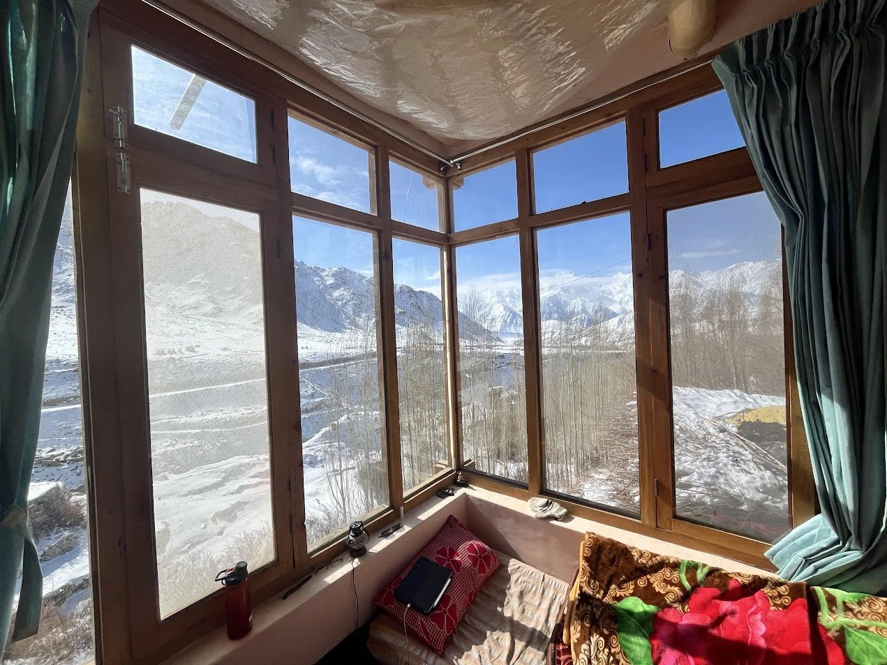 Room with large corner windows showing snow-covered mountains and trees outside, light blue curtains on sides, small bed with patterned blanket and pillow, and a few personal items inside.