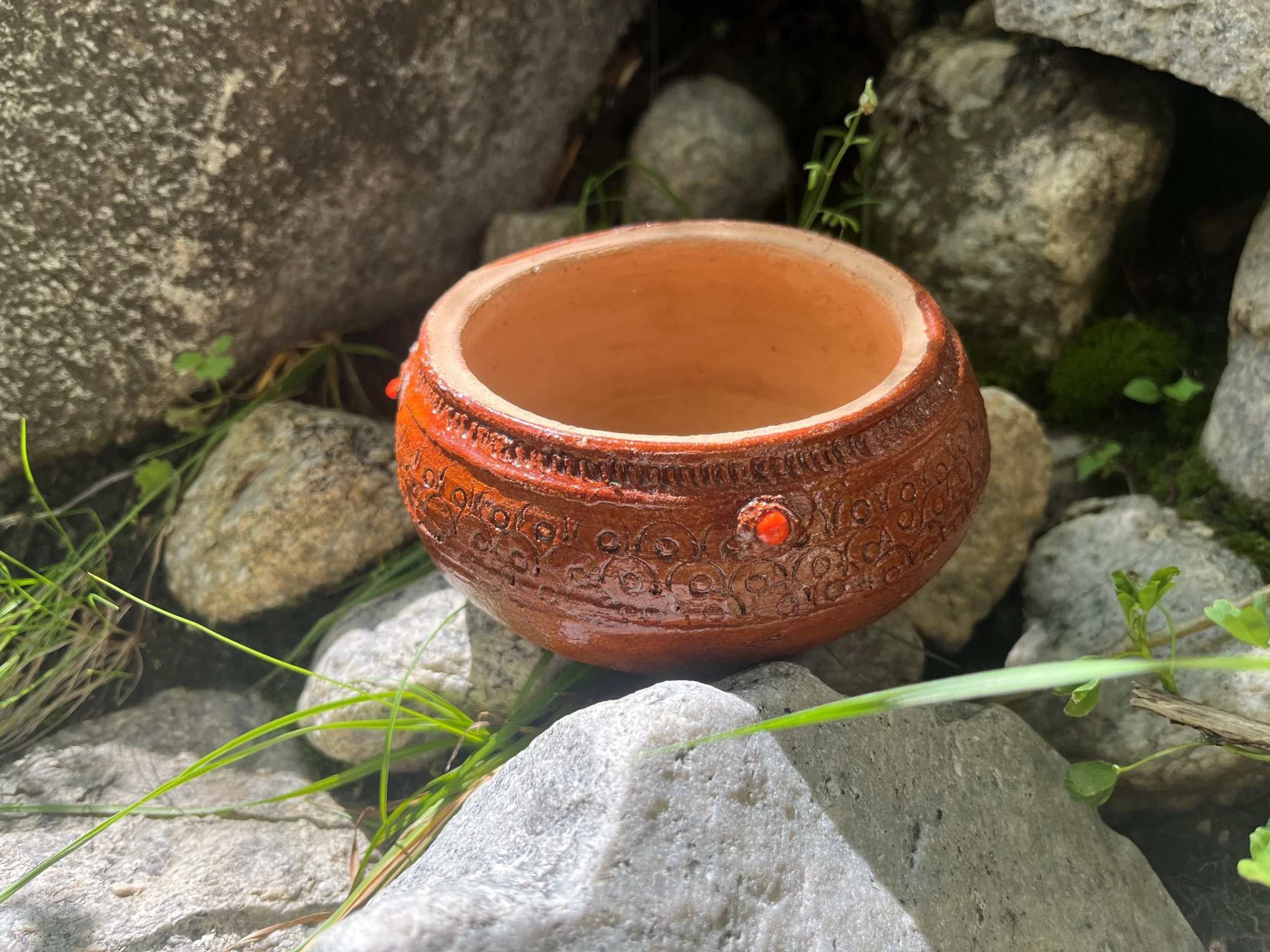 A terracotta pot with decorative patterns, placed among stones and greenery.