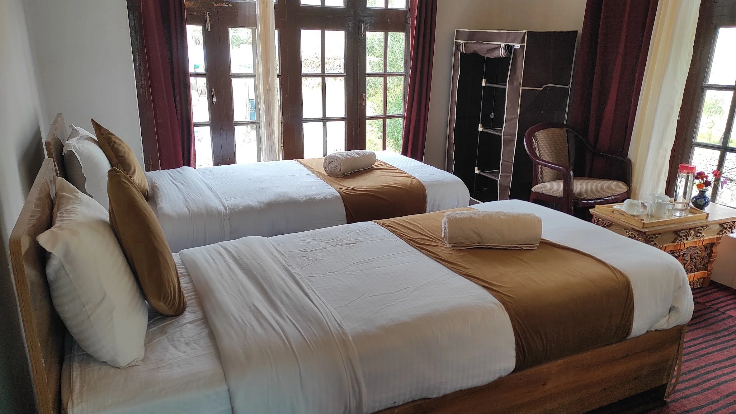 A hotel room with two beds, each with white linens and brown pillows. The beds have rolled towels placed on top. Near the beds, there is a small wooden table with a tray holding cups, a teapot, and a vase with flowers. In the background, large window