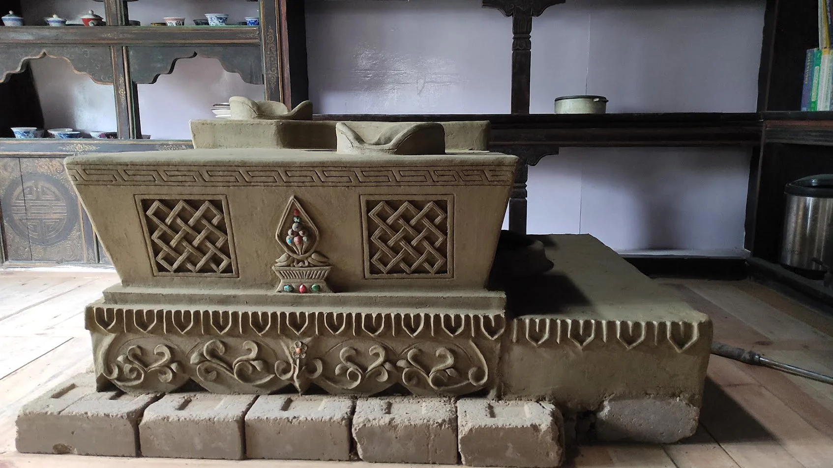 A carved clay stove (thap) with intricate patterns and a bone-like sculpture on top, placed on bricks inside a room with wooden shelves and cups in the background.