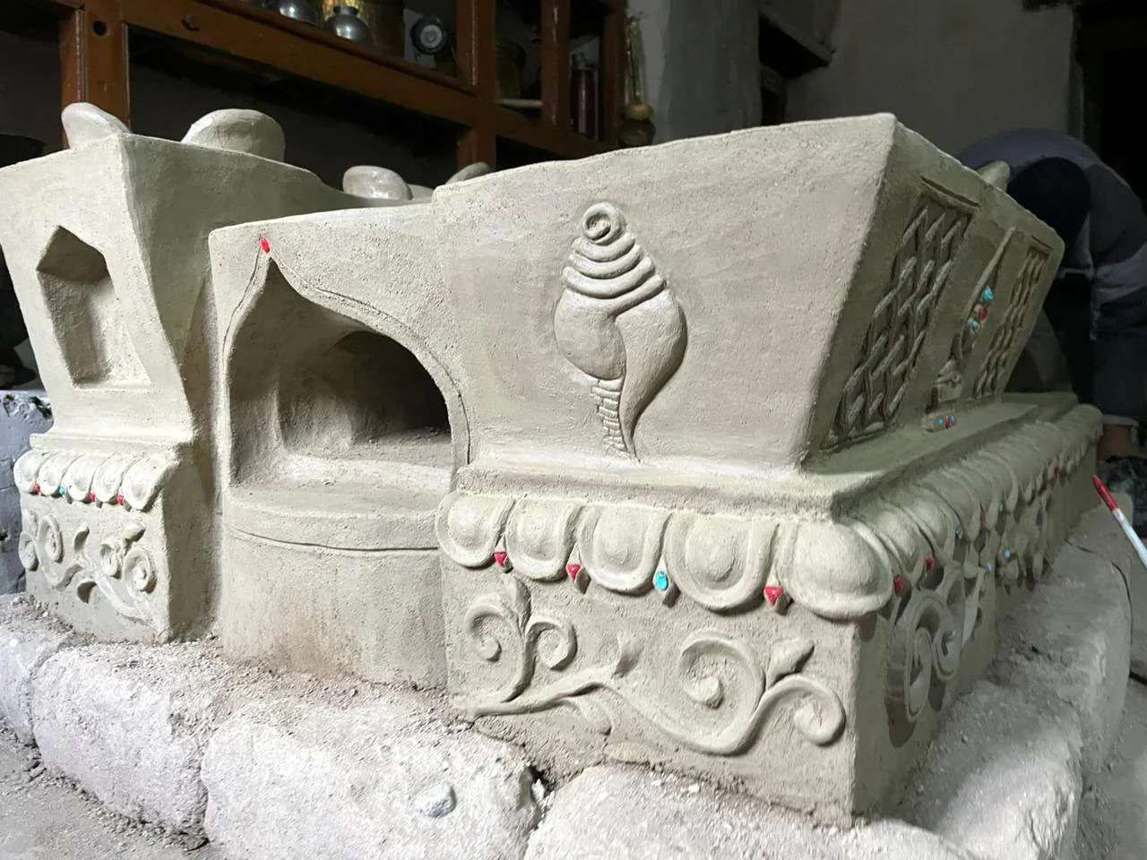 Unfinished decorative stone sculpture with intricate carvings and a shell motif in the center, set on a stone base, with workshop background.
