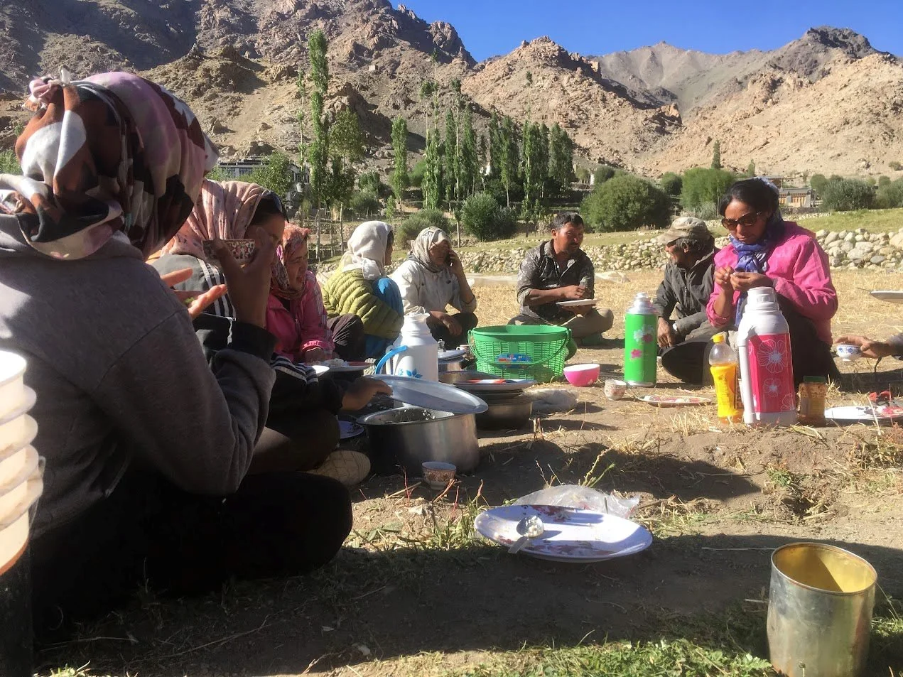 Group of people sitting on the ground outdoors during daytime, with mountains and trees in the background, enjoying a meal with various containers and dishes.