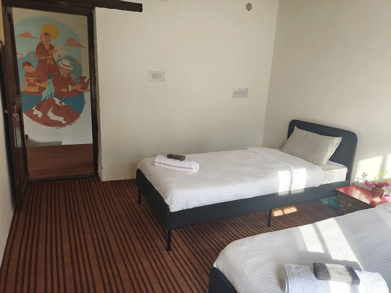 A simple hotel room with two beds, one with a pillow and a rolled towel, and a nightstand with a small potted plant and pink decorative items. The room has a striped carpet and a wall with multiple electrical outlets, with sunlight casting shadows in