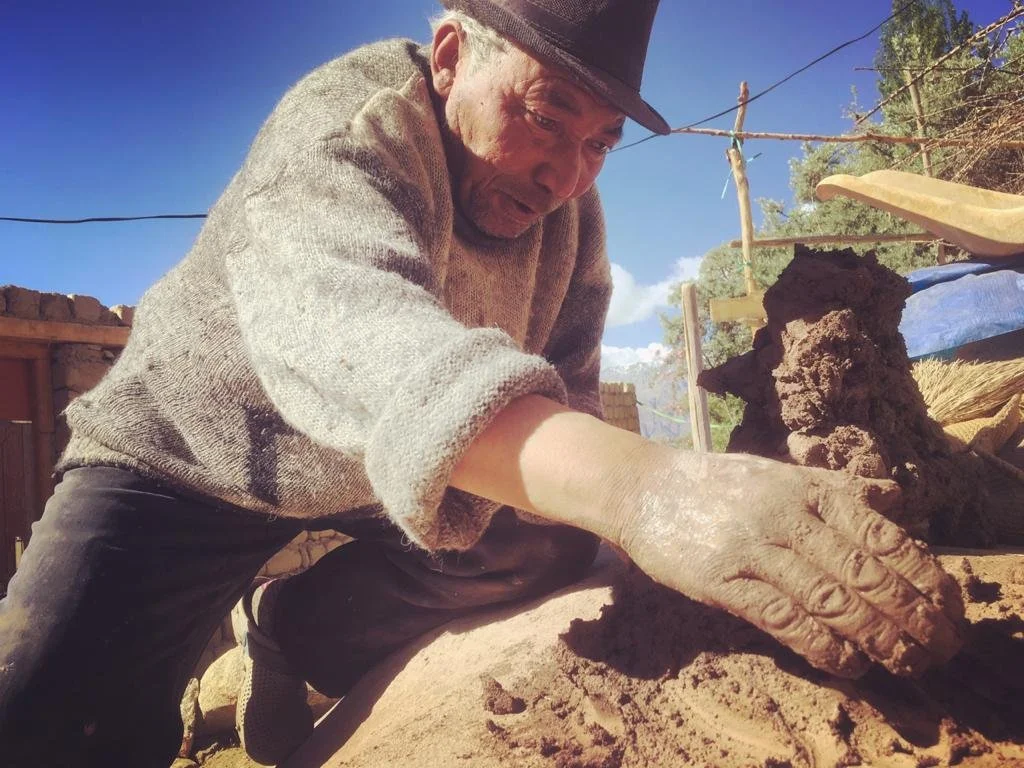 A man with a black hat and beige sweater working with clay outdoors, with a wooden and wire fence, trees, and blue sky in the background.