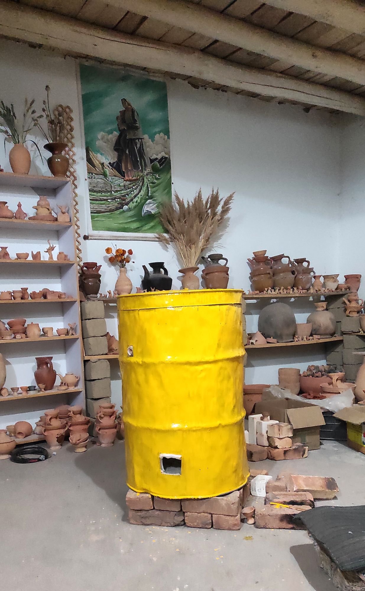 A room with shelves filled with clay pots and small sculptures, a yellow barrel in the center, and a large painting of a woman in the background. The room appears to be a pottery workshop and a yellow kiln.