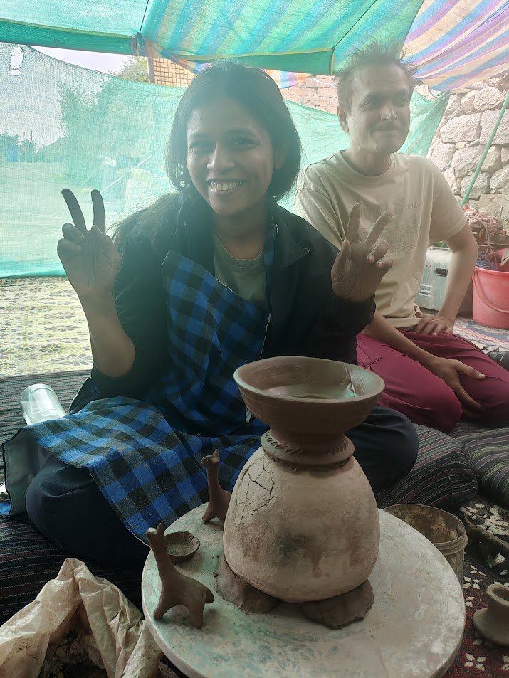 Two people smiling and making peace signs while sitting indoors behind a pottery wheel with unfinished clay pots and small animal figurines, under a colorful tent-like covering.
