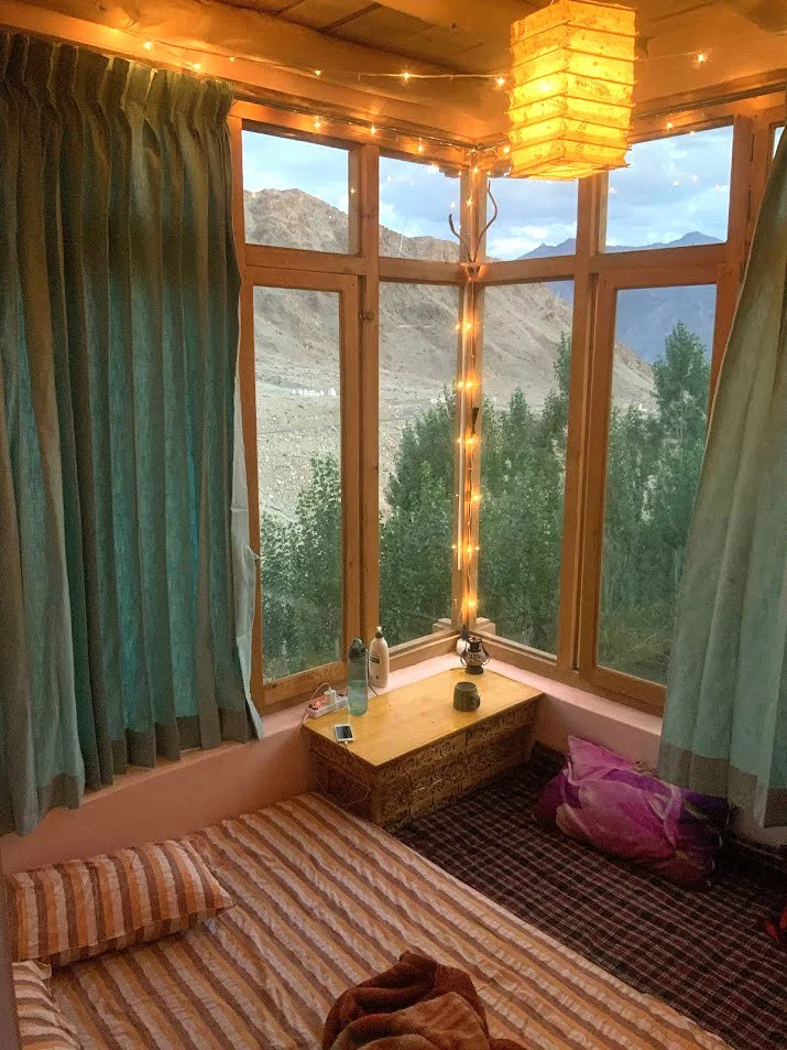 A cozy room with a bed on the floor, a window seat with a pillow, a corner with candles and bottles, green curtains, and string lights, overlooking mountains and trees outside.