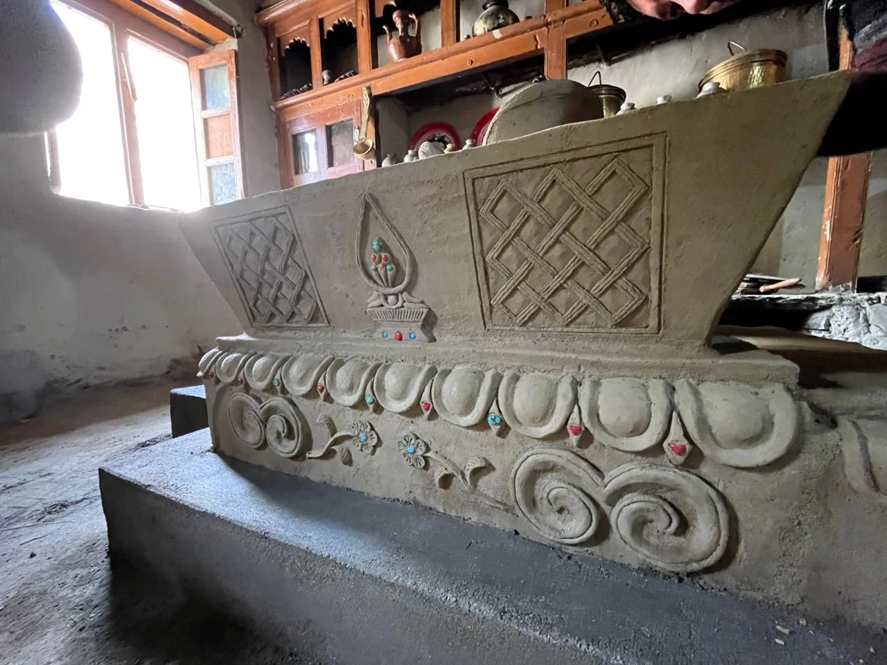 An ornate clay stove (thap) with detailed carvings and inlaid colorful stones. It is situated in a rustic room with wooden shelves and a window in the background, letting in natural light.