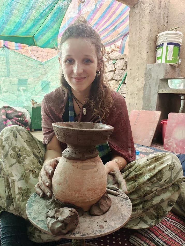 Young woman with long, curly hair and a friendly smile, seated on the ground in a rustic workshop, holding a partially finished clay pot she has crafted. The workshop has a brick wall, colorful tarp overhead, and clay tools and materials around her.