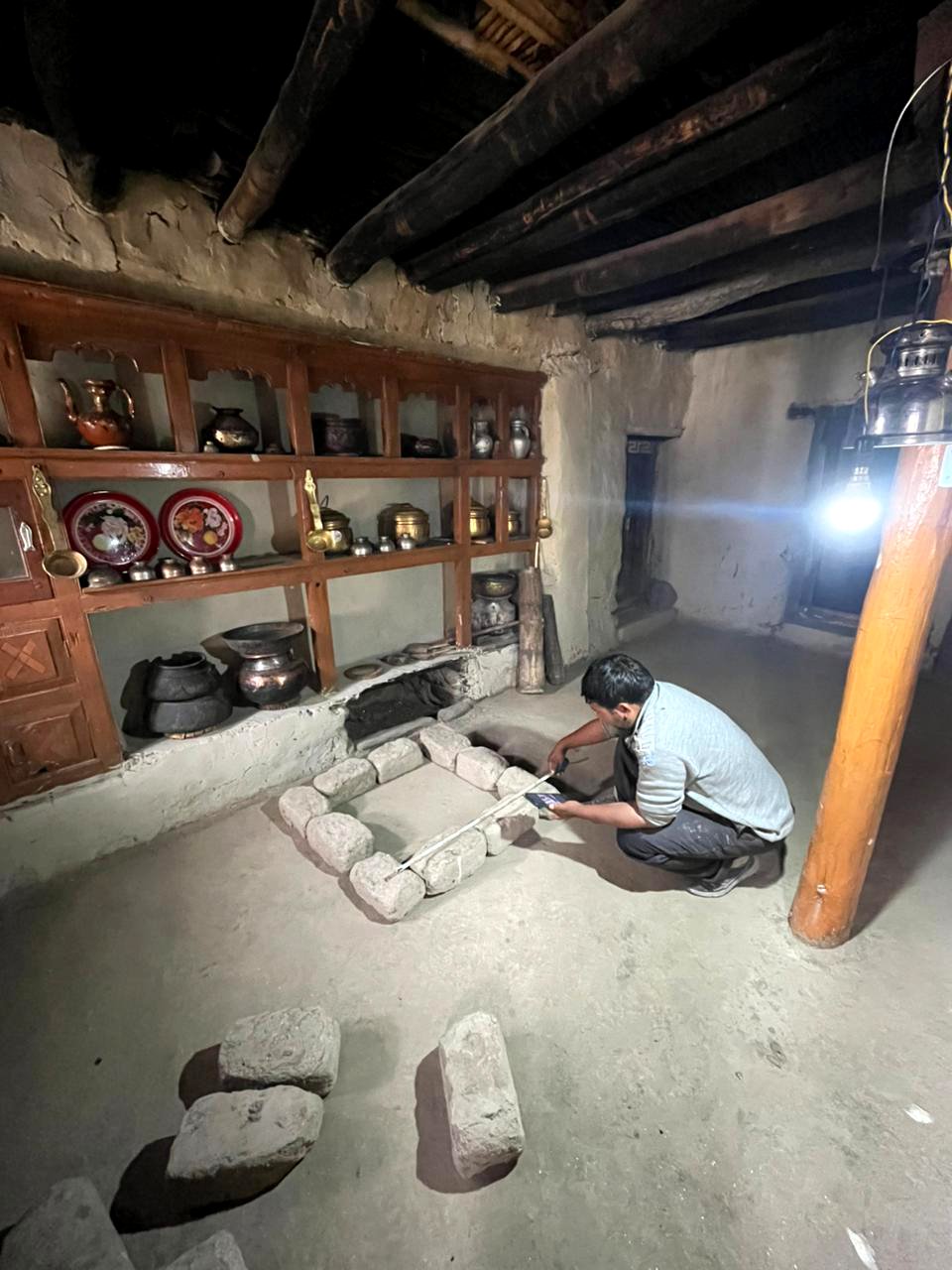A man squats inside a traditional medicine or cultural room, measuring a small stone square with a tape measure. The room has wooden shelves with decorative pottery and metal containers, and a rustic design with exposed wooden ceiling beams and earth