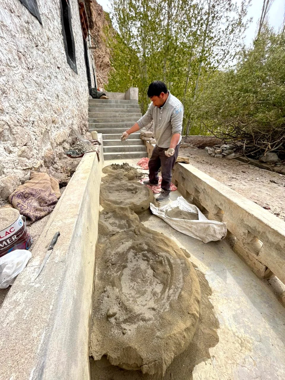 A man working on a preparing  clay outdoors beside a stone wall and a staircase, surrounded by trees.
