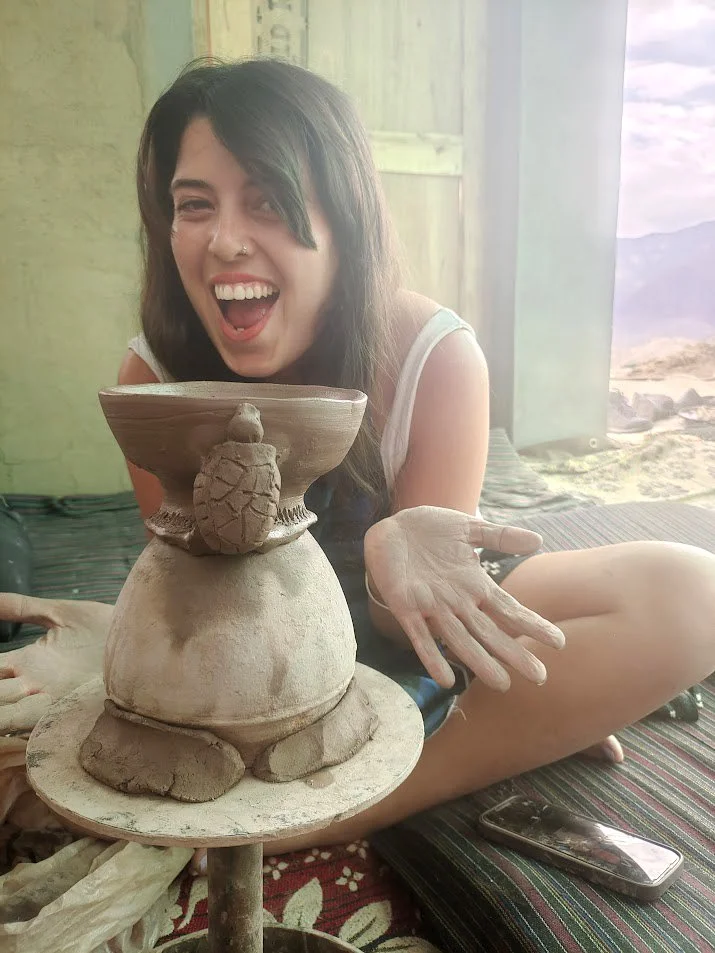 Young woman with dark hair and a nose piercing, sitting cross-legged on a striped mat indoors, smiling and showing her empty hand while displaying a small pottery sculpture of a turtle on a larger clay vessel. A smartphone is near her on the mat, wit